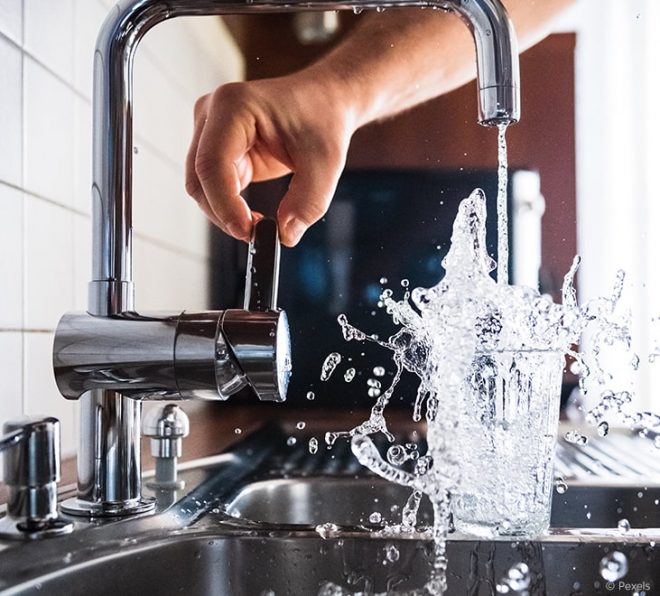Wasserhahn wird über einem Spülbecken geöffnet. Der Wasserverbrauch ist enorm, da das Wasser aus einem überfüllten Glas herausspritzt.