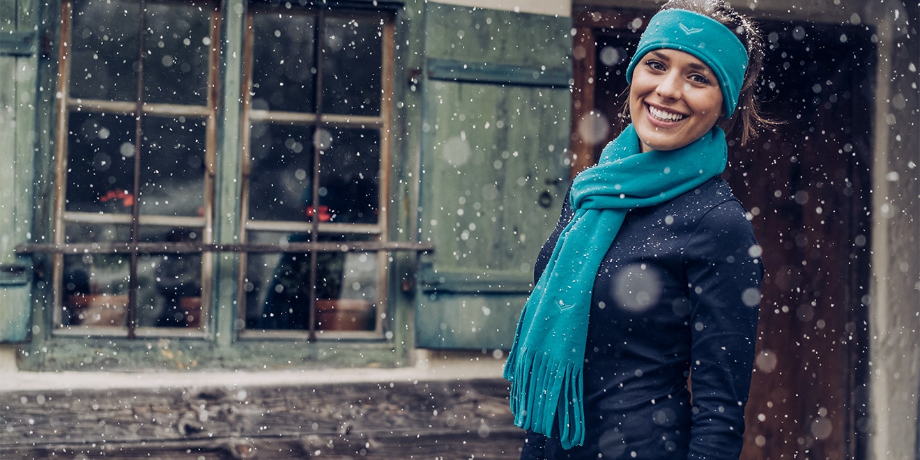 Dame mit Stirnband und Schal in identischen Farben im Schnee vor einer Holzhütte.